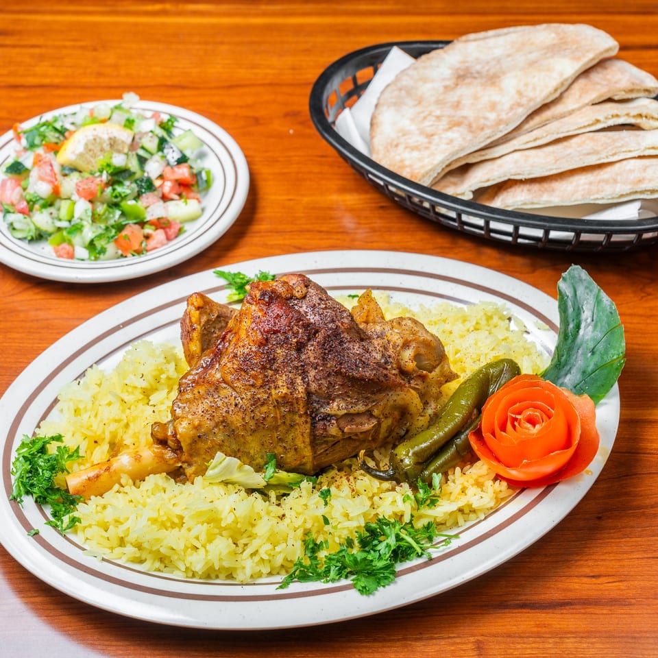 Middle-Eastern lamb shank with yellow rice, green peppers, tomato rose, salad, and pita bread.