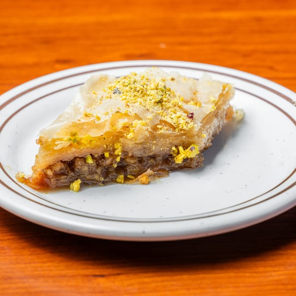 A triangular slice of baklava with flaky pastry, nuts, and honey, topped with crushed pistachios.